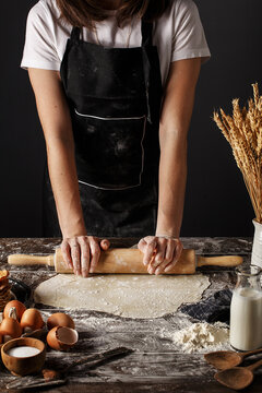 Woman in black apron rolling out dough on wooden table with spilled flour, raw eggs, bottle of milk and wheat spikes. Step by step recipe of cooking dough, bakery products or pastry
