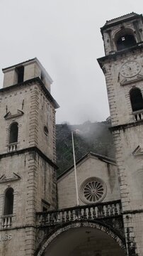 Low-angle view of historic stone cathedral with dual bell towers set against misty mountains on overcast rainy day. The Cathedral of Saint Tryphon in Kotor, Montenegro