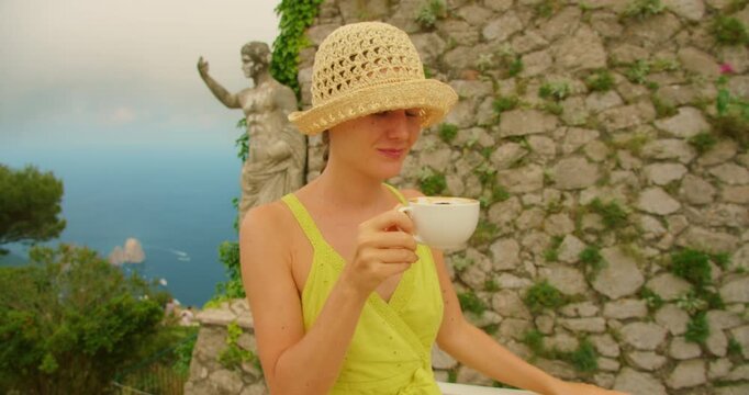 Woman drinking coffee near a stone wall with statue of Emperor Augustus above the sea in Capri Italy