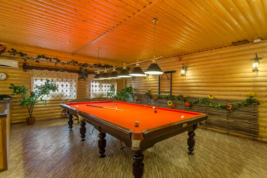 The scene shows a billiard table in a small room with wooden walls. Lights hang above the table. Plants decorate the space and the mood feels inviting for a game in the evening