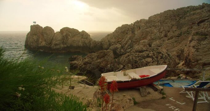 Small boat resting on rocky Capri shore Faro di Punta Carena beach at sunset near rugged Mediterranean coast