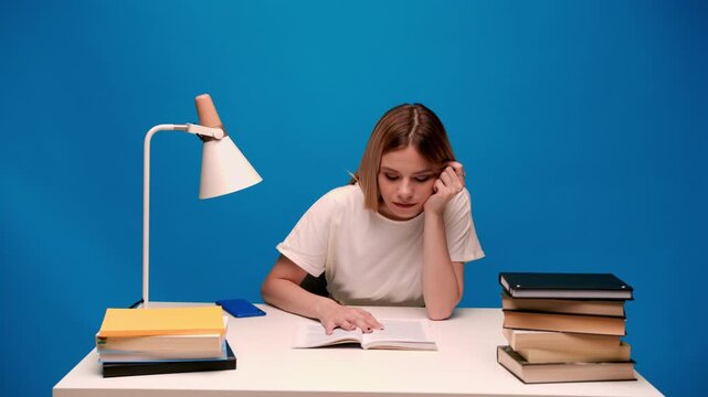 Young female student reading a book and studying at her desk with a stack of books on a blue background
