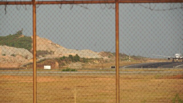 Chain link fence frames a barren landscape. Distant hillside holds loose rock and sparse trees. Lone red structure sits near a paved road. No people or animals are visible
