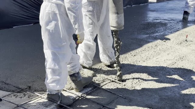 Workers in white suits pour concrete. They use a vibrator to level the mix. Sunlight casts sharp shadows on the ground. Scene shows active construction site. Focus on hands and tools in motion