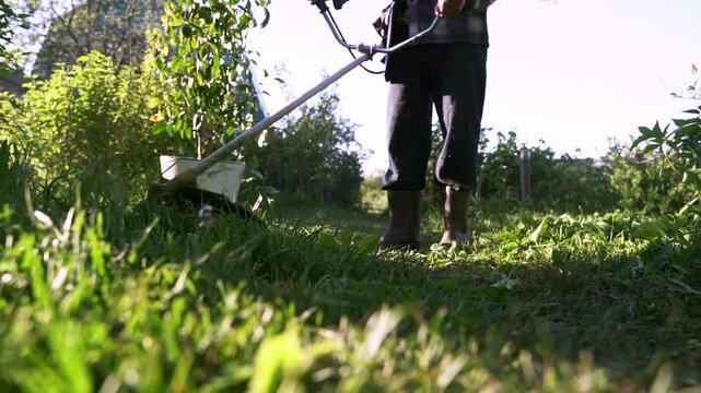 Person pushes a grass trimmer through tall green grass. Sunlight glints off blades and the trimmer handle. Their boots crunch on dry, cut grass. They move slowly, focused on the task