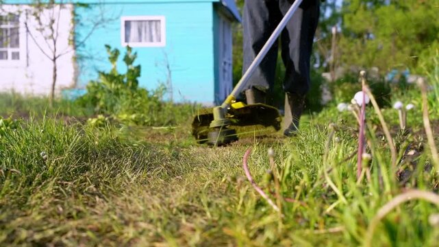 Person mows grass with a handheld tool. Green blades slice through overgrown turf. Sunlight glints off grass and tool. Background shows a bright blue house. Person moves slowly, focused on task