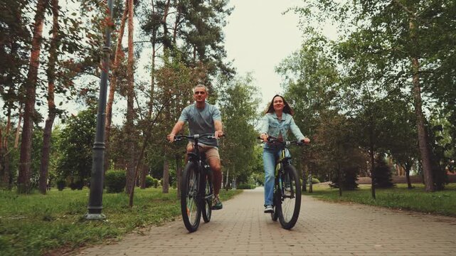 Smiling man and woman ride bicycles along paved path in lush park surrounded by trees, enjoying outdoor activity and leisure time together