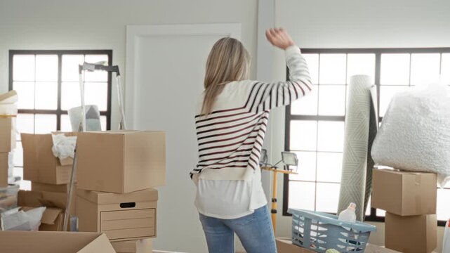 Woman middle age blonde dancing among moving boxes and laundry basket, arm raised gesture in a cluttered living room; joy fresh start.
