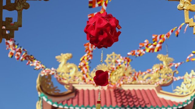 Macro shot of red decorative lantern and flower pendant at Loc Tho Pagoda entrance. Blurred Buddhist flags and golden temple roof on blue sky background. Lunar New Year in Nha Trang, Vietnam.