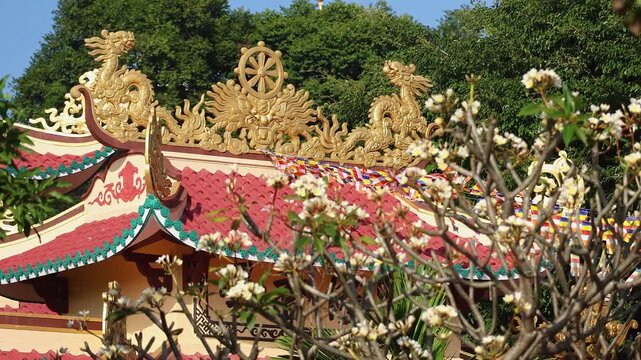 Beautiful view of Loc Tho Pagoda roof architecture with golden dragon statues and dharma wheel. Traditional Buddhist temple decorated with flags against green trees and blue sky. Nha Trang, Vietnam.