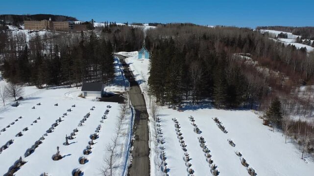 Winter aerial view of the snow-covered Lac-au-Saumon cemetery leading to the small blue Saint-Joseph Oratory and the historic convent of the Servants of Our Lady Queen of the Clergy in the background.