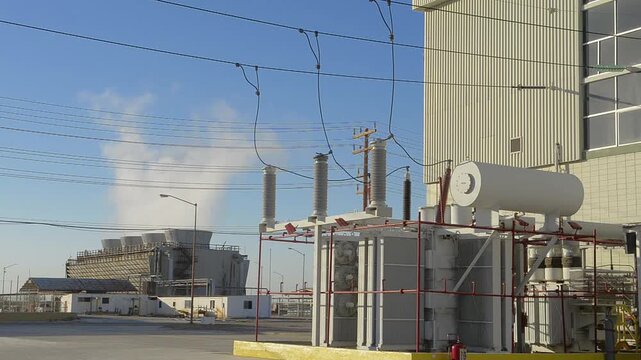 Transformador principal de una planta generadora de energia electrica conectada a una subestacion para red electrica de transmision, con una torre de enfriamineto en el fondo.