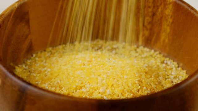 Yellow corn grain flour for polenta porridge falling down into the wooden bowl close up