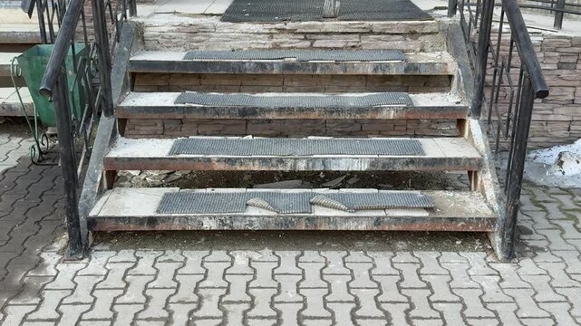 Worn antiskid coating on steps at pickup entrance, rusted metal treads with peeling paint and grime, uneven concrete pavers, damp.