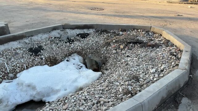 Snow crushed stone around sewer pipe, sunlit urban curb and asphalt, melting patches and scattered pebbles, exposed drain hole.
