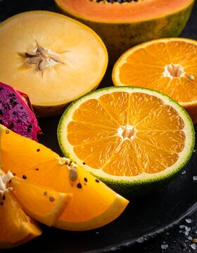 Sliced tropical fruits, including orange, papaya, dragon fruit, sapote, on a dark plate. Fresh and vibrant