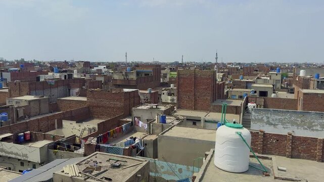 Cinematic panning rooftop view of a densely populated middle class residential neighborhood in Faisalabad, Punjab, Pakistan, featuring brick houses, water tanks, and mosque minarets