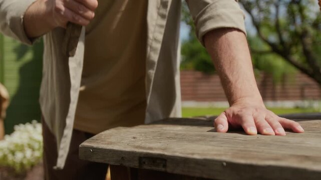 Medium midsection slowmo shot of unrecognizable rural man beating on nail with hammer while repairing wooden table or chair outdoors in garden, busy with chores in village household