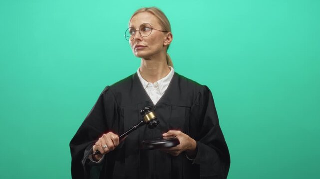 Woman judge in black robe raises gavel with right hand over wooden sounding block in studio green screen, wearing glasses and white collar; authority justice.