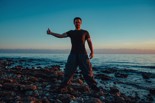 Man standing on rocky shore at sunset giving thumbs up