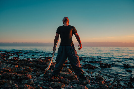 Man holding wooden stick on rocky beach at sunset
