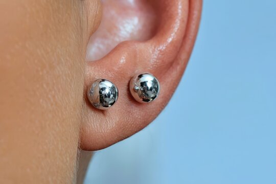 Close-up of a human earlobe adorned with two shiny silver spherical stud earrings against a soft blue background.