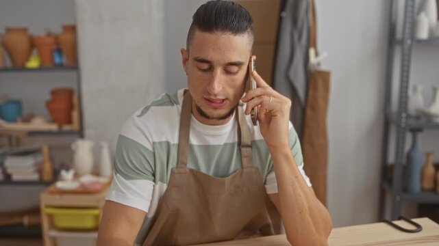 Man in apron holds phone to ear while gesturing and inspecting a pottery tool on the table in a studio; thoughtful craft focus.