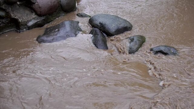 Panning view of muddy brown floodwater rushing over river rocks in Indonesia, illustrating the environmental effects of heavy monsoon rains and upstream soil erosion in Southeast Asia.