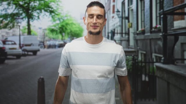 Man with palms open and slightly raised, wearing striped t shirt, standing on a city street with parked vans and iron railings; invitation calm.