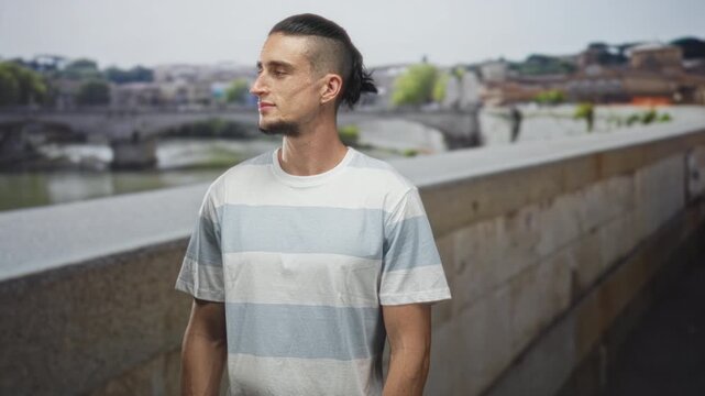 Man in striped tshirt standing on street by stone bridge facing camera with arms at sides and neutral gaze; contemplation.