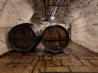 Two weathered wooden wine barrels rest in a rustic stone cellar, surrounded by rough-hewn walls and cobblestone flooring. Dim lighting highlights the aged wood and stone textures, creating an authenti © stanhert