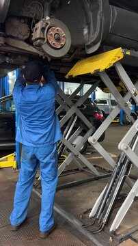Car mechanic working under lifted vehicle in garage workshop during auto repair service. Brake maintenance, suspension inspection and professional vehicle service in automotive garage.