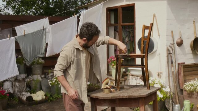 Medium full shot of crafty Caucasian village man repairing antique furniture in backyard, carefully painting vintage wooden chair, laundry hanging on lines and blooming flowers in background