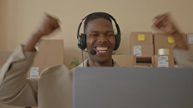 Man raising fists and smiling while wearing headset at laptop with stacked packages and shipping labels in building; celebration success.
