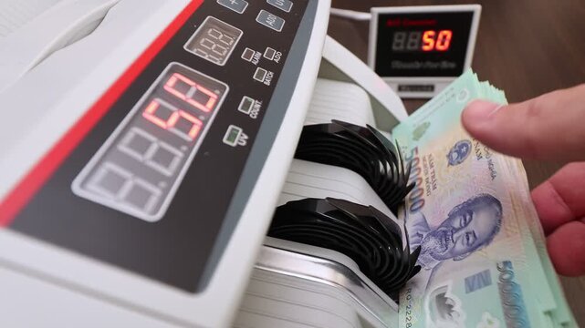 Closeup shot of banknote counting machine with Vietnamese Dong money (VND). Automatic currency counter equipment processing paper bill with Ho Chi Minh portrait vignette. Finance industry in Vietnam.
