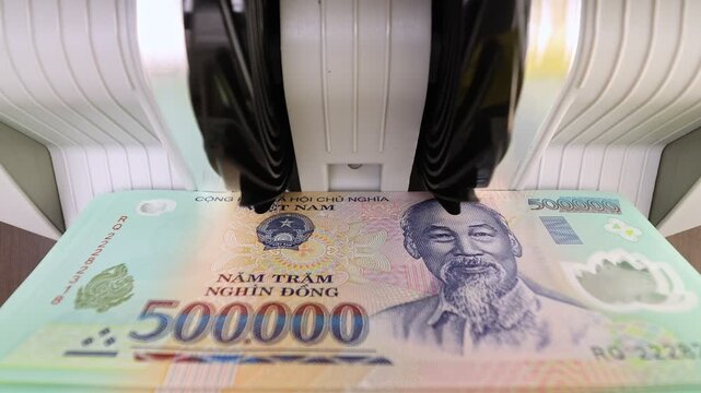 Closeup shot of banknote counting machine with Vietnamese Dong money (VND). Automatic currency counter equipment processing paper bill with Ho Chi Minh portrait vignette. Finance industry in Vietnam.