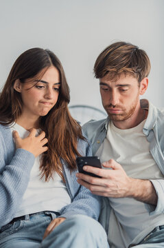 A young couple sits closely on a couch, both looking worried and upset as they view messages on a smartphone, conveying distrust, jealousy and strained communication in a domestic setting.