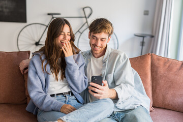 Fototapeta premium Young couple sits close on a sofa in a cozy living room, smiling and laughing as they look at a smartphone together, sharing a light, intimate and joyful moment at home.