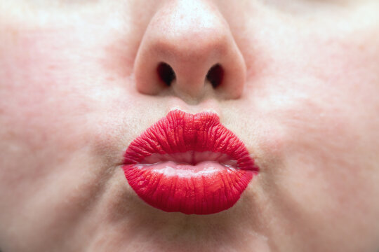 Extreme close-up of a woman's face with red lipstick puckered in a kiss. Macro portrait showing lips and nose against soft skin background. Beauty, romance and cosmetics concept.