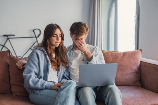 A young couple sits close on a sofa looking worried as they review finances on a laptop, showing anxiety, stress and concern about bills, budgeting and online banking at home.