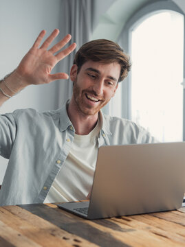 A smiling man waves at his laptop during a friendly video call in a bright home office, conveying remote work, connection, casual meeting and positive communication.
