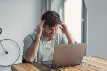A frustrated young man rubs his temples and grimaces while working on a laptop at a wooden table in...