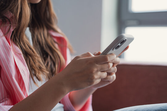 A young woman sits on a sofa using her smartphone, focused on typing and scrolling. The close-up of her hands and device conveys casual communication, connectivity, and everyday digital lifestyle.