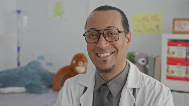 Man doctor wearing stethoscope and glasses smiling beside a toy filled pediatric exam bed in a clinic room; calm reassurance.