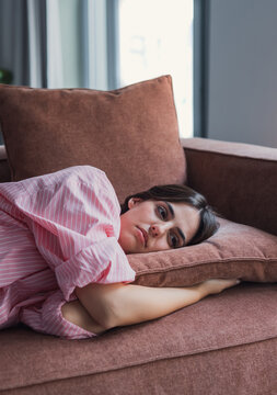 A young woman in a pink striped shirt lies on a rust-colored sofa, resting her head on a cushion while appearing tired, pensive and lonely in a quiet domestic living room.