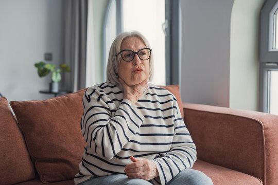 Senior woman with sore throat sitting on couch at home showing discomfort and pain