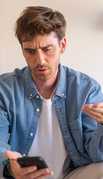A young man in a denim shirt looks frustrated and confused while reading his mobile phone at home, conveying stress, worry and concern about a message or unexpected news.