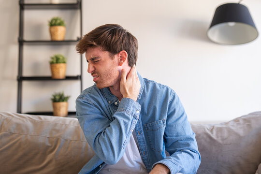 Young man sitting on a sofa at home grimacing and holding his neck in pain, suggesting neck strain, discomfort or injury; casual interior and natural light convey everyday health concern.