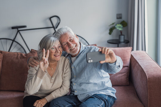 A smiling senior couple sit close on a plush sofa, taking a smartphone selfie while the woman flashes a peace sign. Warmhearted moment conveys love, companionship and aging happily at home.
