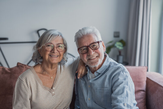 Smiling senior couple sit closely on a sofa in a bright living room, wearing glasses and casual clothes, sharing a warm, affectionate and relaxed moment together at home.
